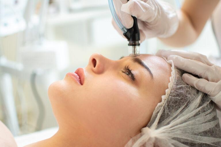 Close up of therapist's hands in protective gloves holding hydrafacial tool and performing anti aging progress to beautiful young woman forehead at beauty salon Image description